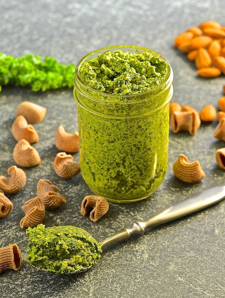 Kale Almond Vegan Pesto in jar with a spoon of pesto to the side and scattered pasta shapes