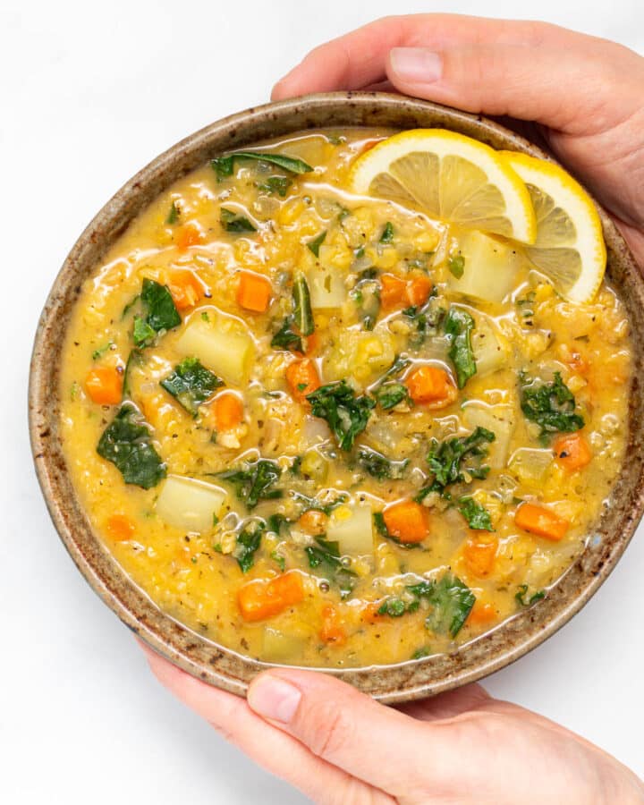 A bowl of lemony red lentil soup with a lemon slice garnish being held by 2 hands