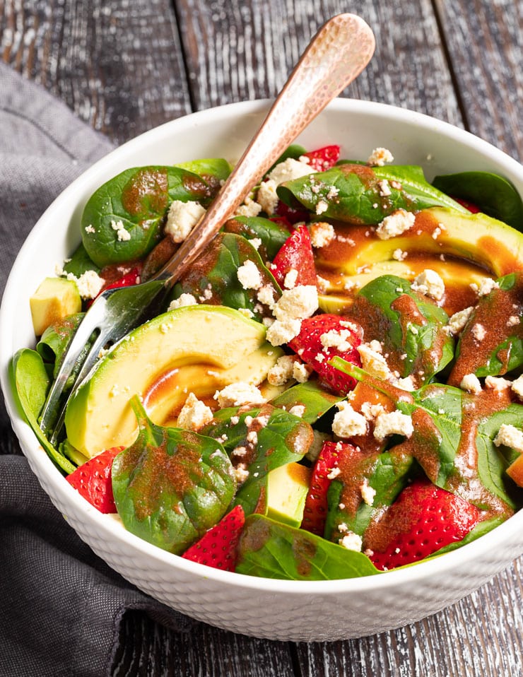 a bowl of strawberry spinach salad with avocado and vegan feta, drizzled in vinaigrette