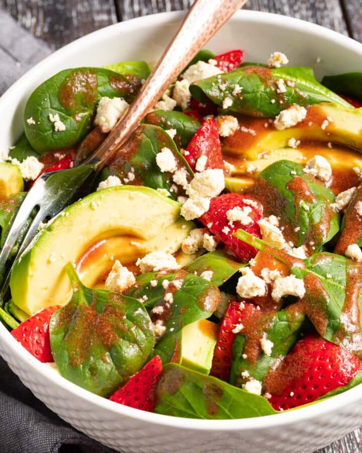 a bowl of strawberry spinach salad with avocado and vegan feta drizzled in strawberry vinaigrette