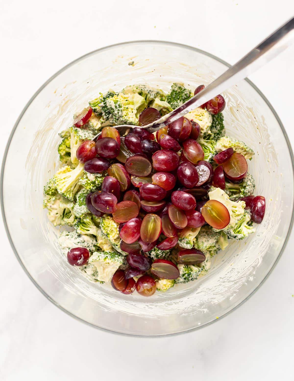 a bowl of broccoli tossed in creamy dressing with red grapes on top