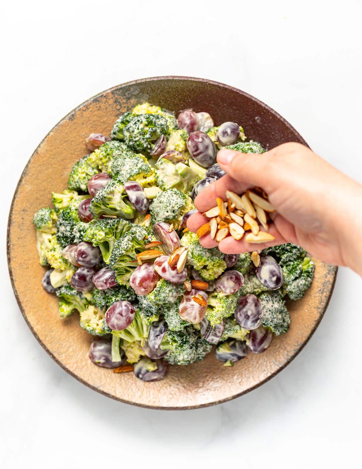 a plate full of vegan broccoli salad with a hand sprinkling over slivered almonds 