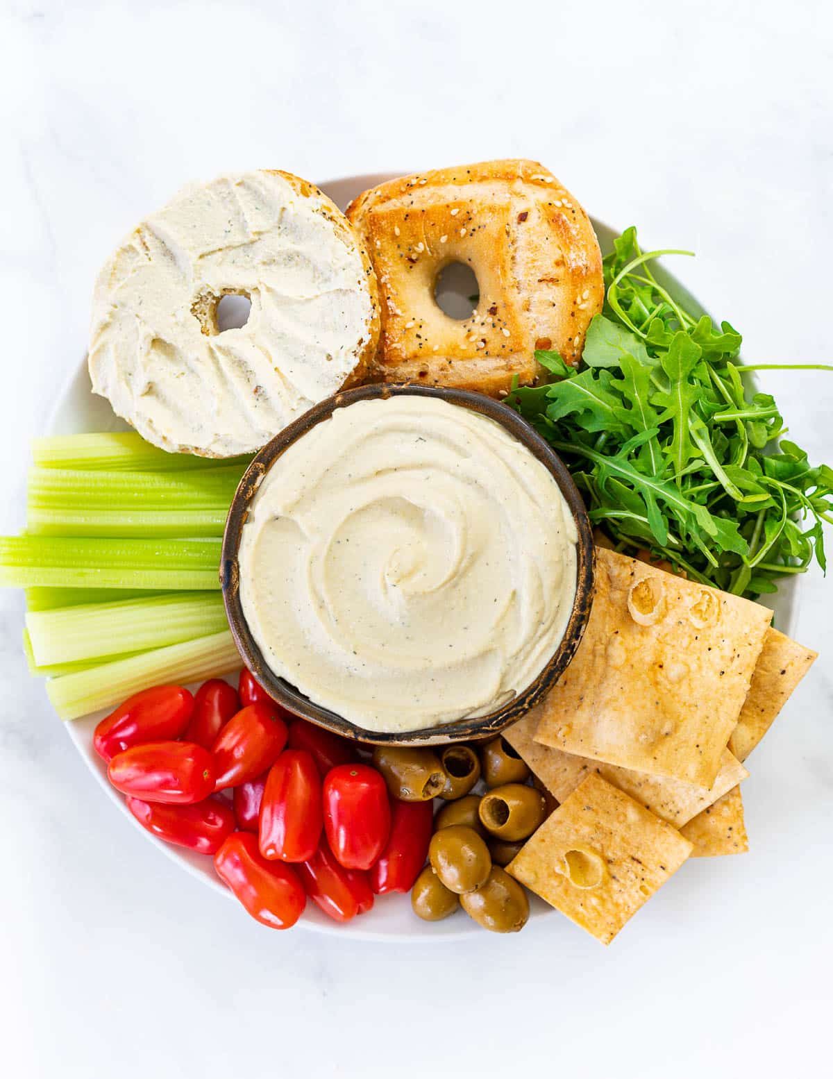 a platter with a bowl of vegan cream cheese, bagels, crackers and vegetables