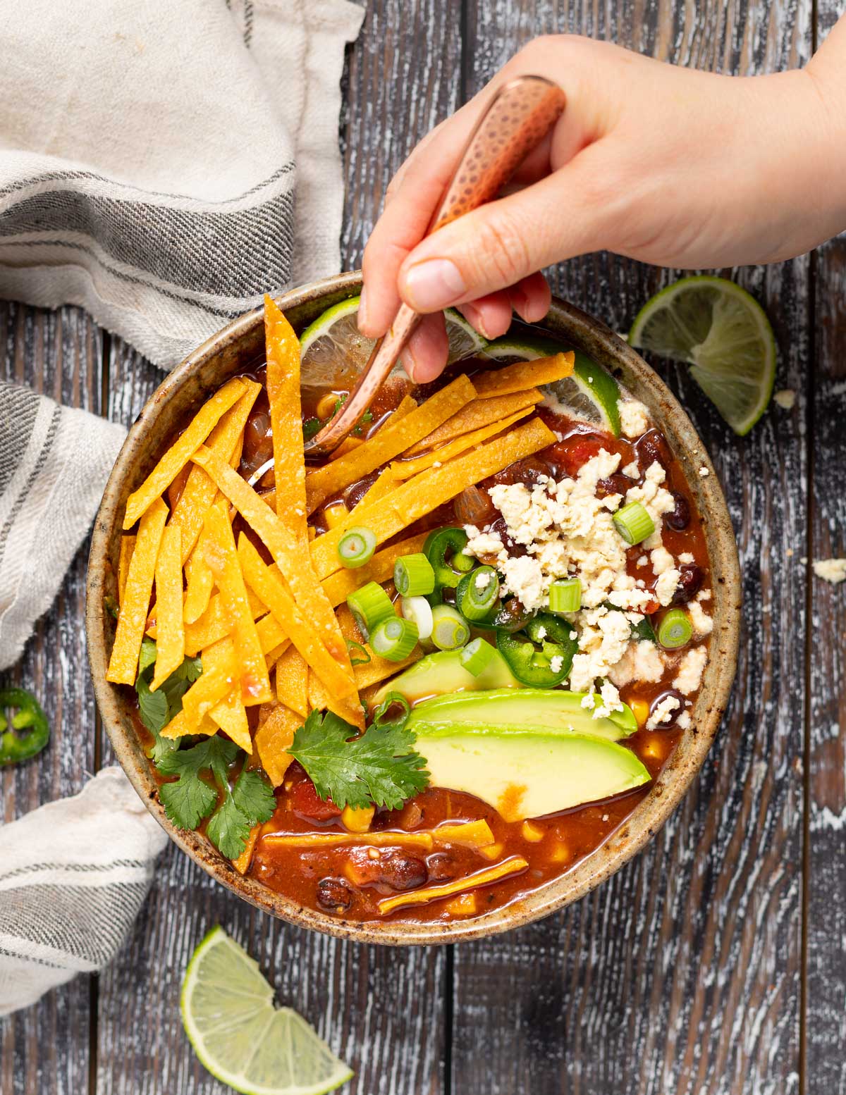a spoon dogging into a bowl ot vegan tortilla soup covered in lots of topping slike avocado, jalapeno, vegan feta cheese and cilantro 